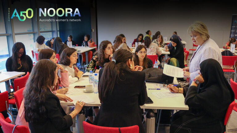 A photo from Aurora50's NOORA women's network (with logo) from a meet-up on 9 January 2025. The workshop focused on social capital and two male allies were invited to attend. In this photo, seven women sit at a table as Prof Dame Heather McGregor of Heriot-Watt University Dubai talks to them directly.